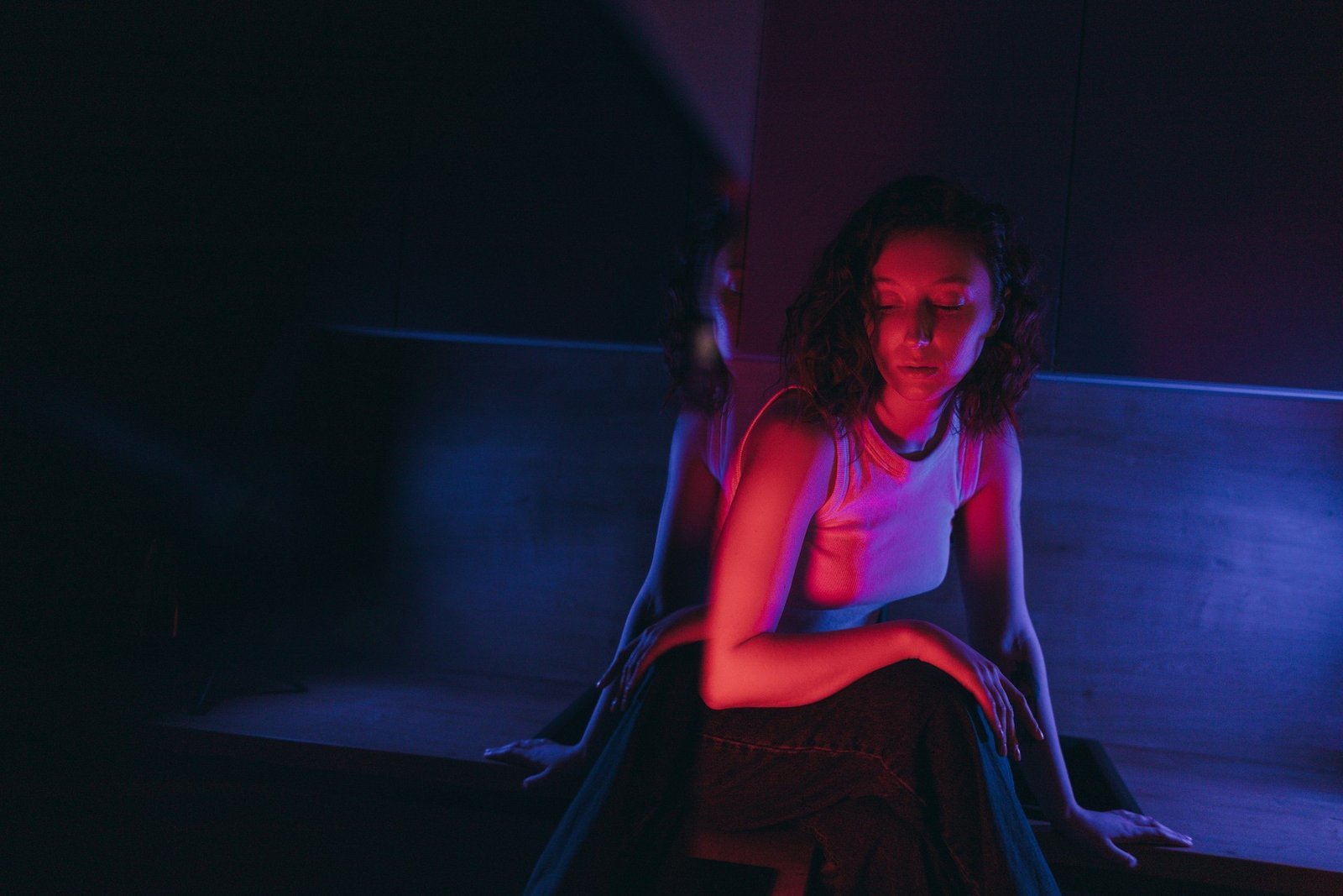 Person seated in a dimly lit room under blue and magenta light, reflecting quietly