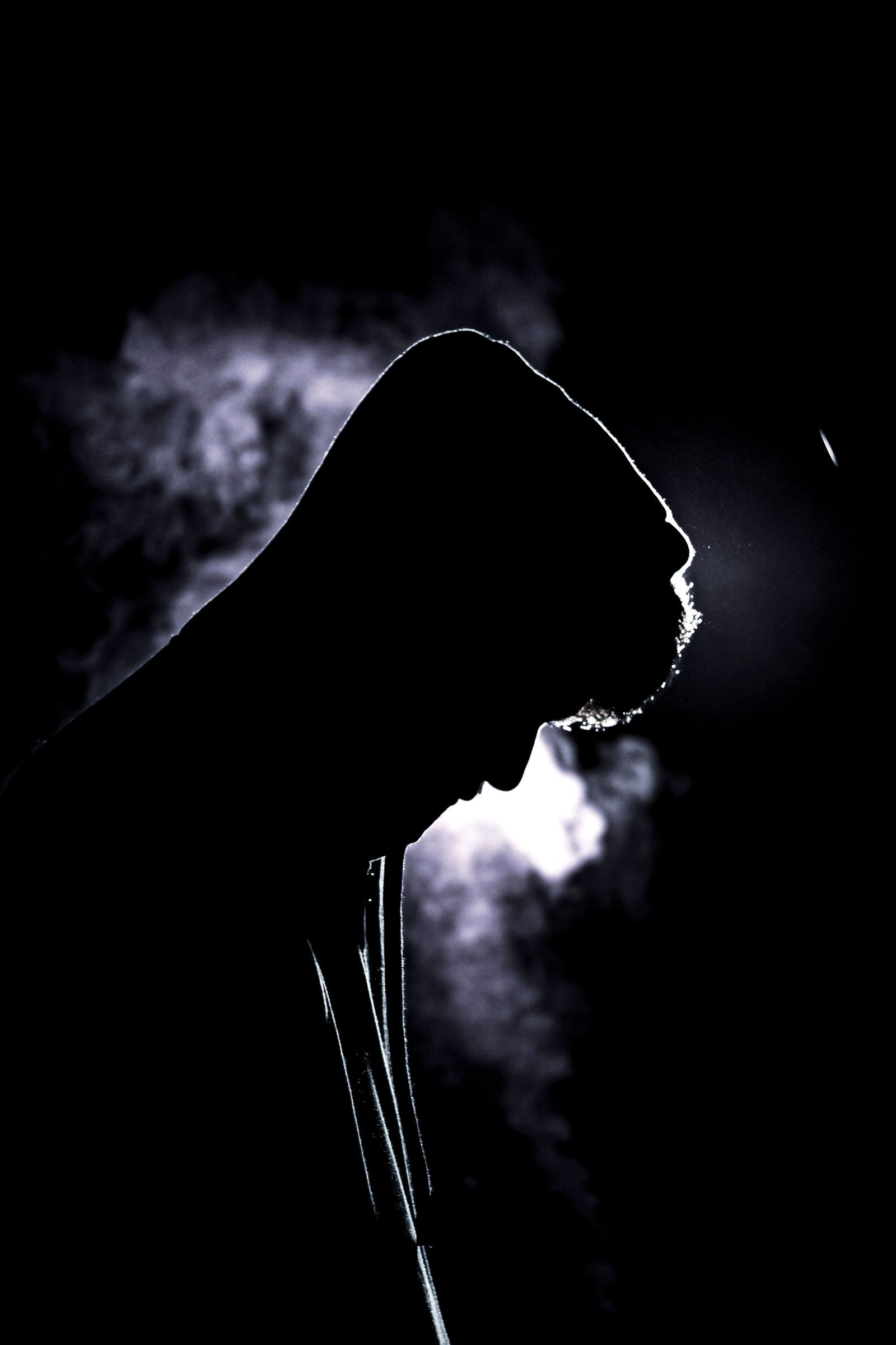 High-contrast silhouette of a person wearing a hood, softly lit against a dark background